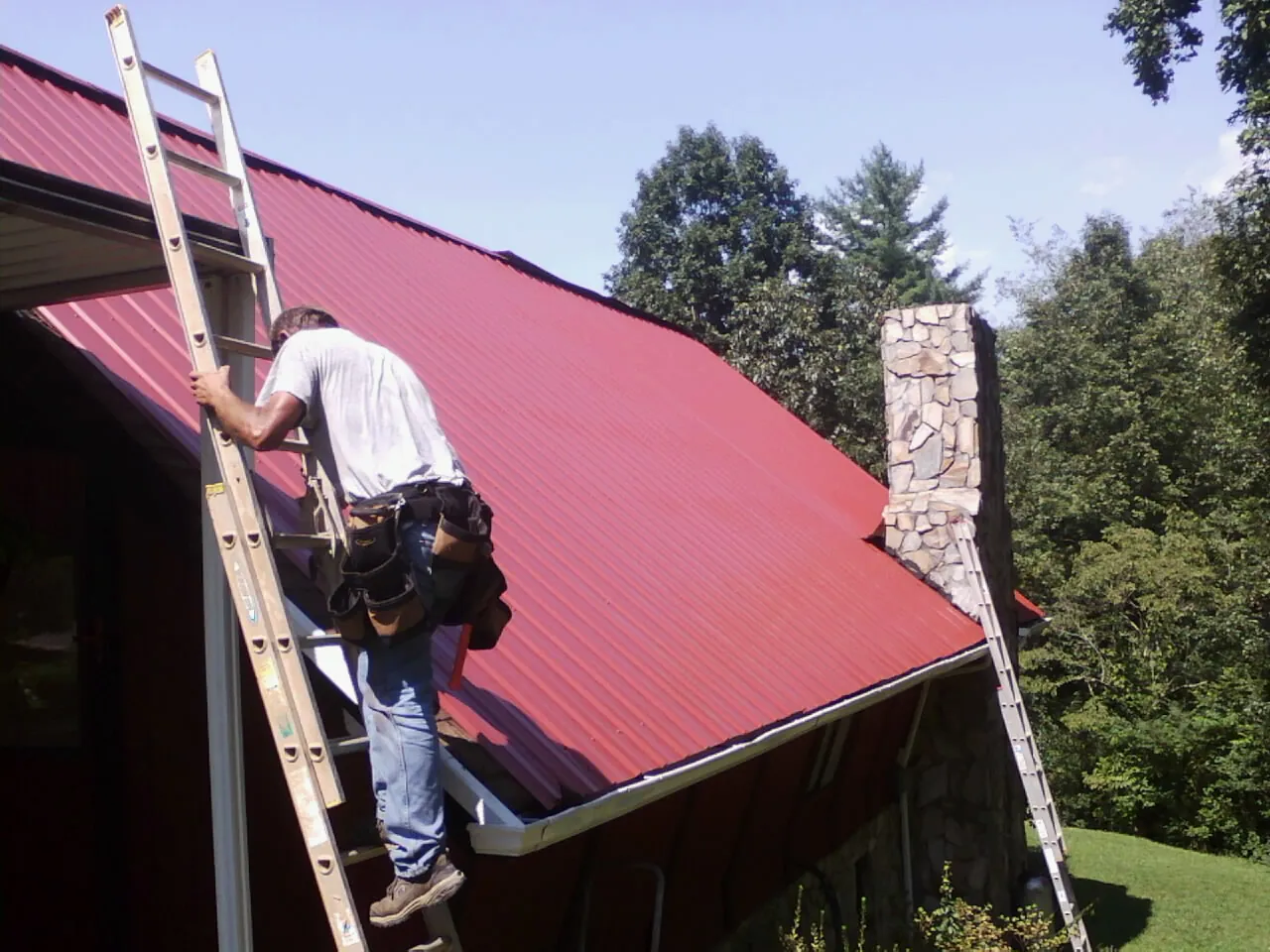 Roofer on a ladder beside a red metal roof and stone chimney — Soffit & Fascia Repair by Roofing Brothers in River Edge